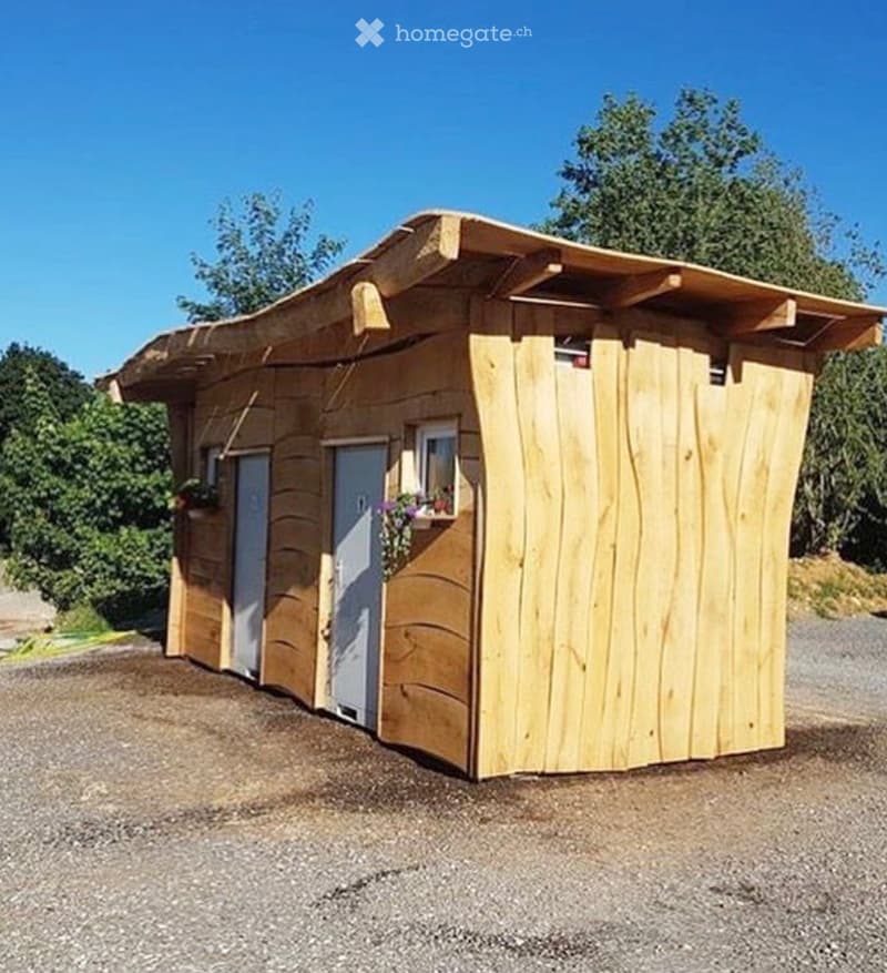 A small wooden cabin with a sloped roof, two doors, and a window with a flower box, situated on a gravel surface with trees in the background.