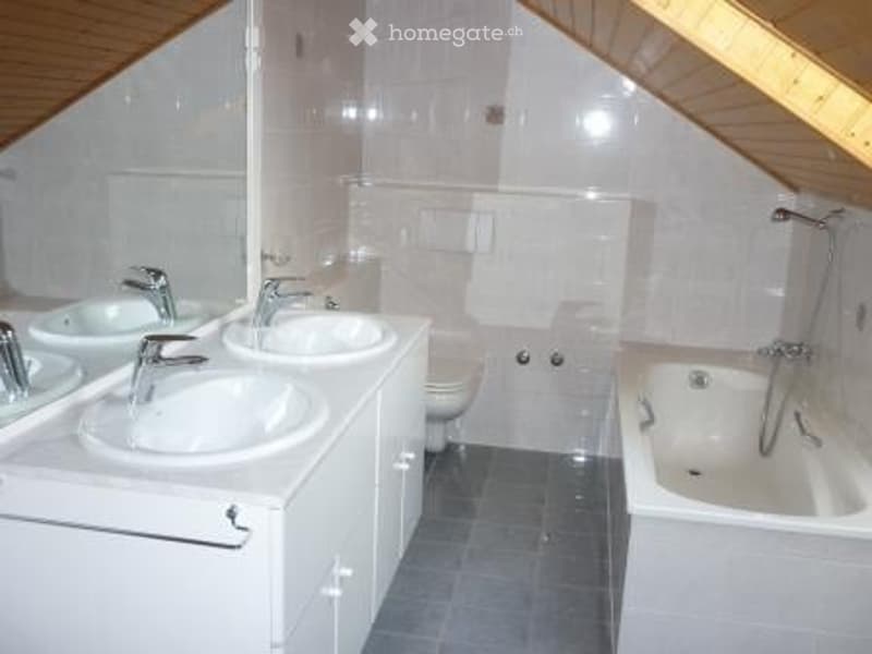 Modern bathroom with two sinks, a bathtub, and a toilet. The room features white tiles and a wooden slanted ceiling.