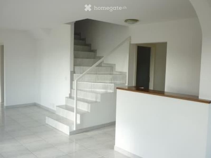 Bright, open hallway with white walls, tiled floor, and a staircase leading upwards. The staircase features a white handrail and gray steps.