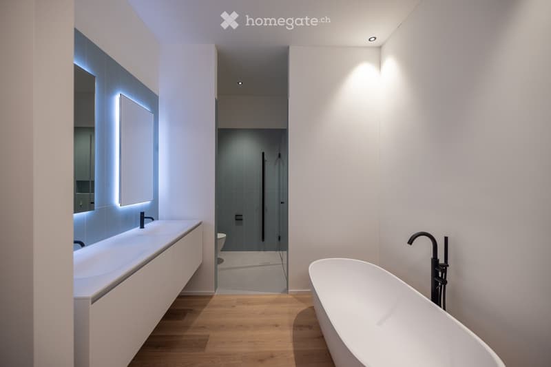 Modern bathroom with a freestanding bathtub, a sleek sink with a modern faucet, and a large mirror with integrated lighting. The space features light wooden flooring and recessed ceiling lighting.