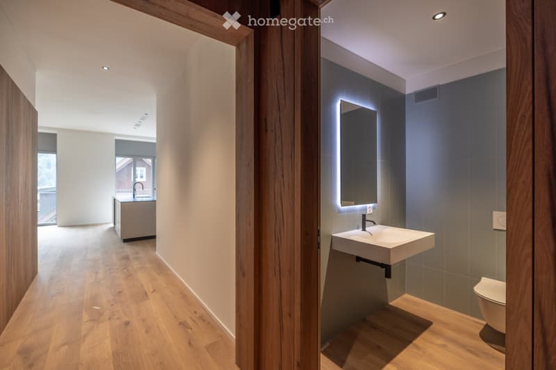 Modern bathroom with wooden flooring, a floating sink, a large illuminated mirror, and a toilet. The space is bright with recessed lighting and sleek, contemporary design.