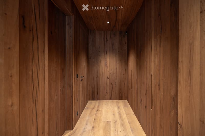 A narrow wooden hallway with polished wooden walls and ceiling, featuring recessed lighting and subtle wood grain patterns.