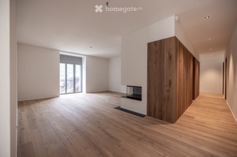 Empty room with light wooden flooring, white walls, recessed lighting, and a fireplace with a glass enclosure. A large window with a sliding door leads to a balcony.