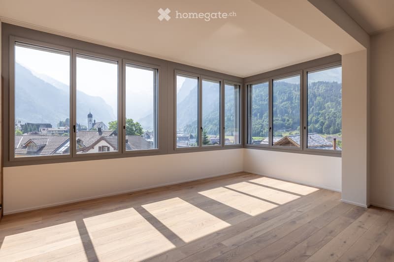 Empty room with wooden flooring and large windows offering a scenic mountain and village view.
