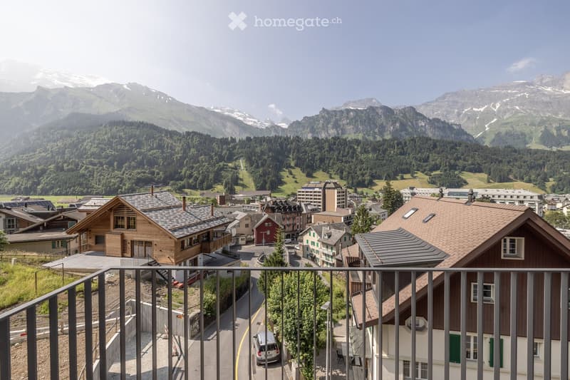 Balcony overlooking a picturesque town with traditional wooden houses, surrounded by lush green forests and snow-capped mountains in the background.