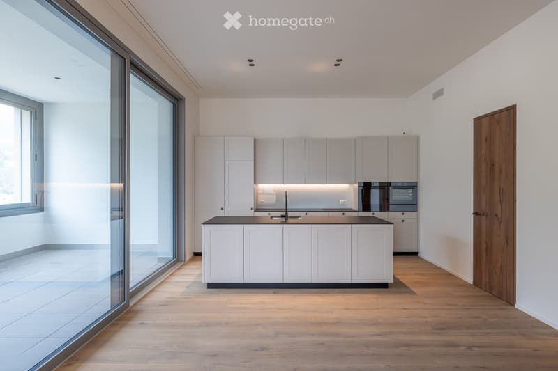 Modern kitchen with white cabinets, a central island, and built-in appliances, featuring large windows and wooden flooring.