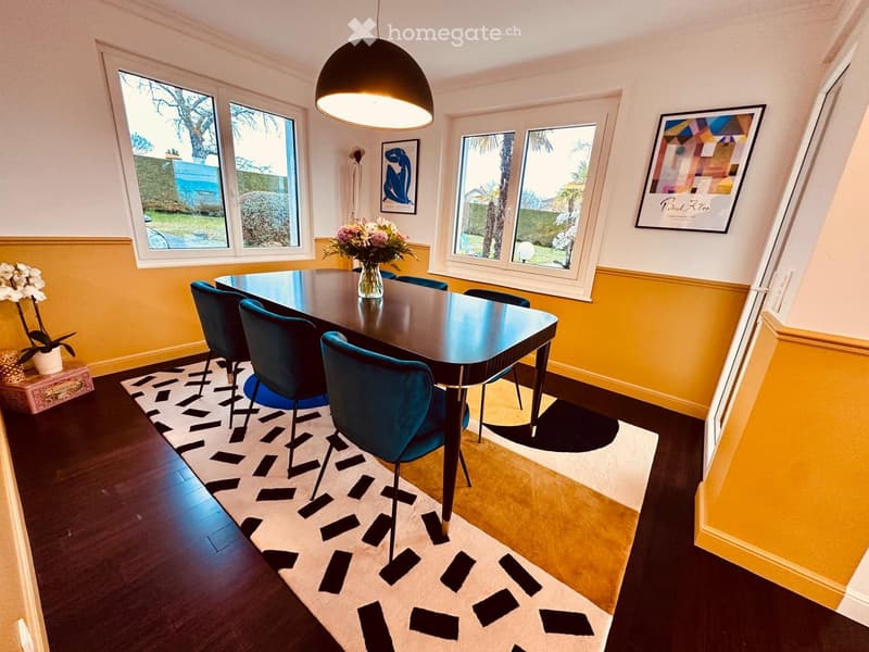 Dining room with a dark wooden dining table, six teal chairs, a vase with flowers, a patterned rug, and artwork on the walls.
