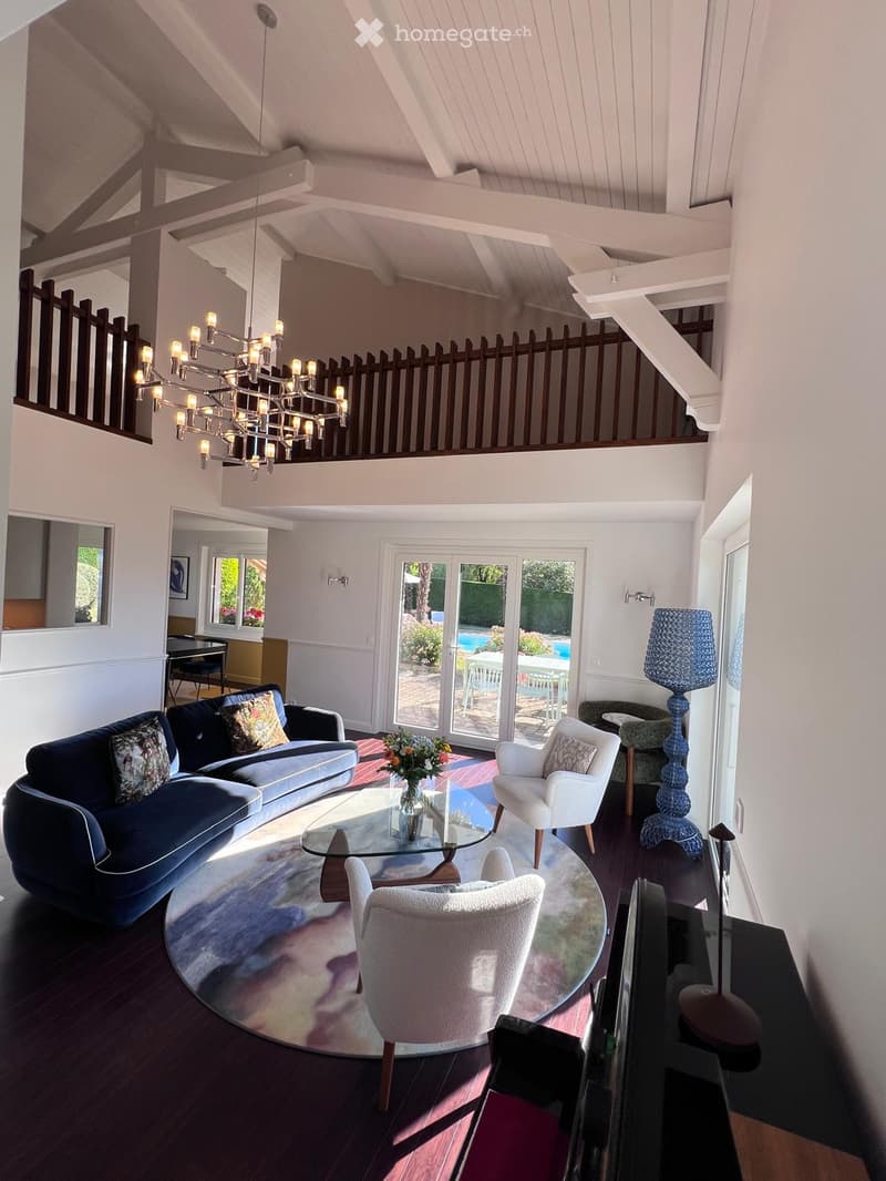 Living room with a dark blue sofa, glass coffee table, white armchairs, a large round rug, and a blue table lamp. The room features exposed wooden beams and a chandelier hanging from the ceiling.