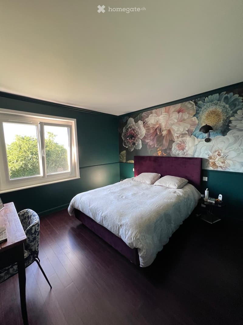 Bedroom with a large bed, nightstand, chair, and table. The walls are painted in dark green with a floral mural above the bed. The room has wooden flooring and large windows letting in natural light.
