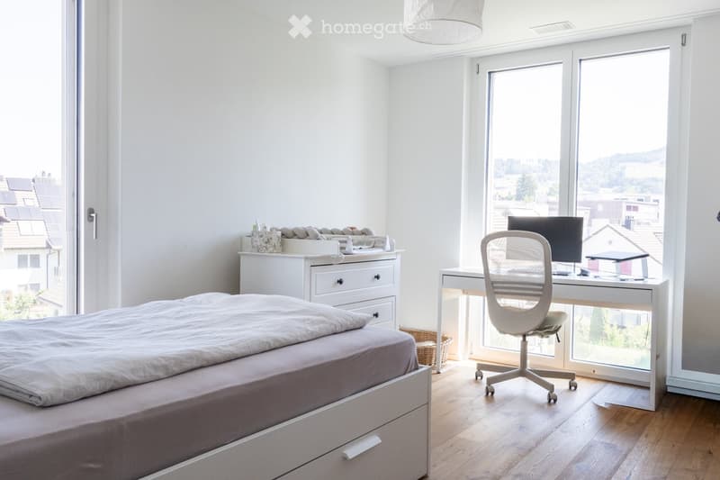 Modern bedroom with a large bed, a desk with a computer and chair, a drawer unit, and a wicker basket. Large windows offer a scenic view.