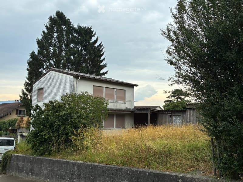 A two-story house with white siding, a pitched roof, and a small balcony on the upper level. The house is surrounded by trees and has a grassy area in front.
