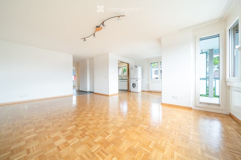Empty room with wooden herringbone flooring, white walls, large windows, and a washing machine in the background.