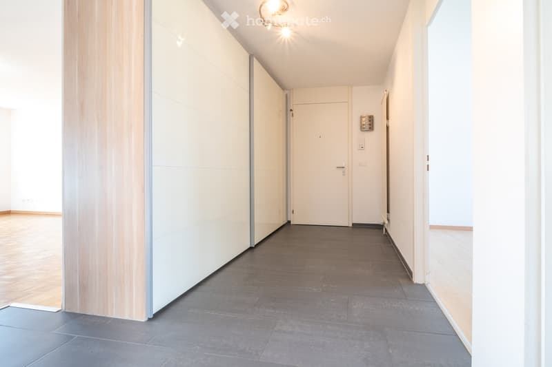 Modern hallway with grey tiled floor, white walls, and wooden paneling on one side. Features a closed door and recessed lighting fixtures.