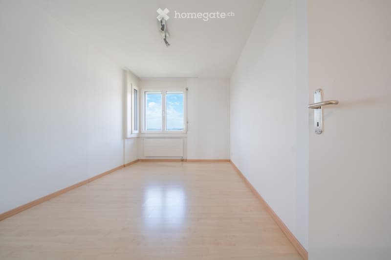 Empty room with light wooden flooring, white walls, and large windows letting in natural light. The room has a simple, clean design with a door on the right side.