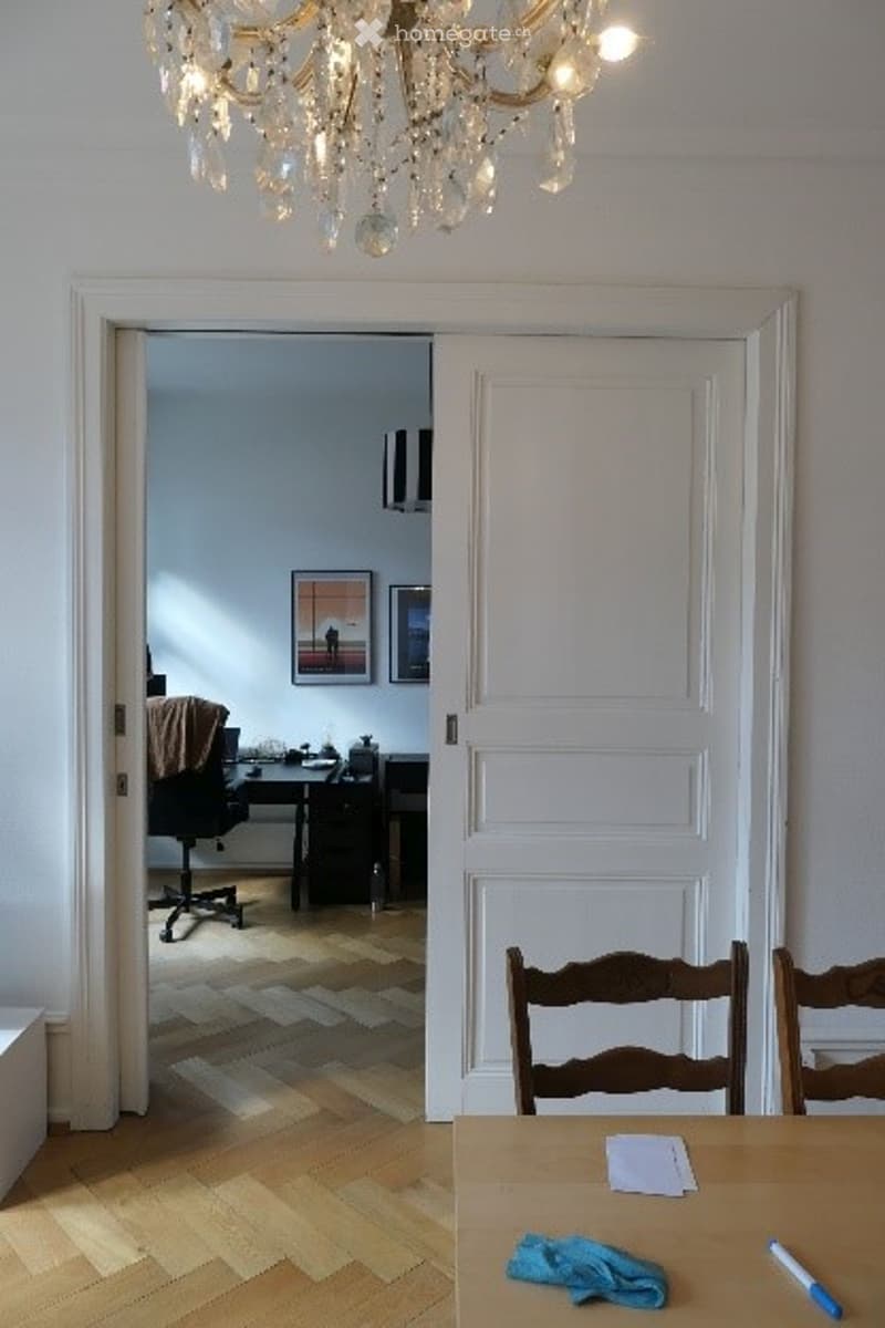 A well-lit room with wooden flooring, a chandelier, a table with chairs, and an open doorway leading to a study area with a desk, chair, and laptop.