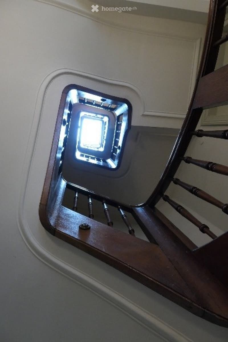 Spiral staircase with wooden handrails and a central light fixture, viewed from below.