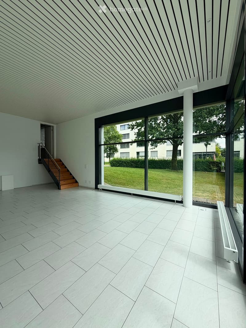 Bright, minimalist room with large glass windows, white tiled floor, and a wooden staircase leading upwards.