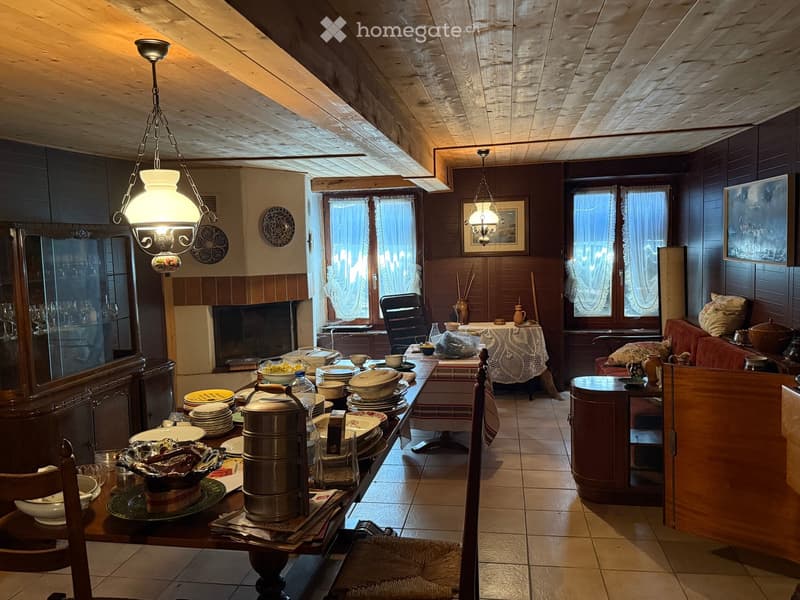 A cozy, rustic living and dining area with wooden beams, a fireplace, a large dining table set with dishes and utensils, a sofa, and a chandelier.