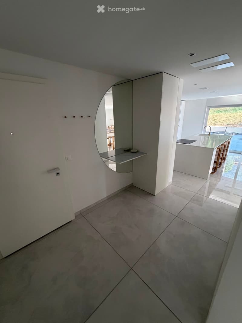 Empty hallway with tiled floor, white walls, and a curved mirror on the wall. The hallway leads to a modern kitchen area visible in the background.