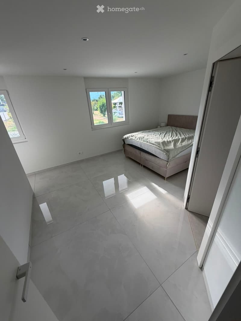Modern bedroom with a large bed, white walls, and large windows allowing natural light to fill the space. The floor is made of polished concrete tiles.