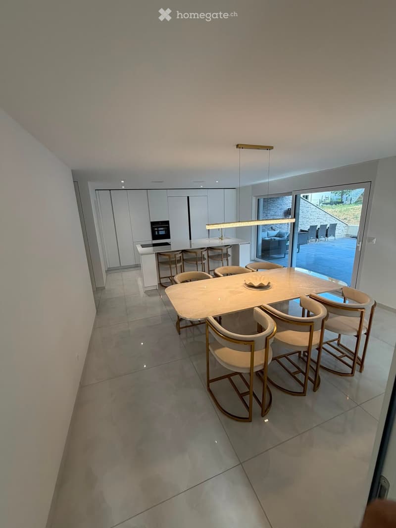 Modern dining area with a large white dining table surrounded by six beige chairs with gold accents, a kitchen island with bar stools, and built-in cabinets with an oven.