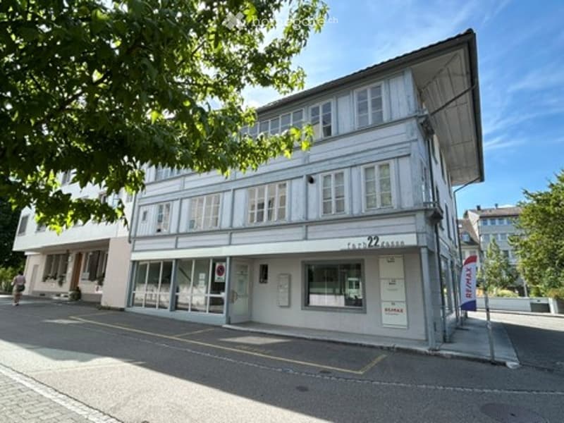 Two-story building with white facade, multiple windows, and a storefront on the ground floor. The building has a modern design with a flat roof and is surrounded by trees.