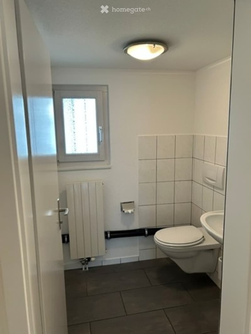 Small bathroom with white tiled walls, a toilet, a sink, a radiator, and a window providing natural light.