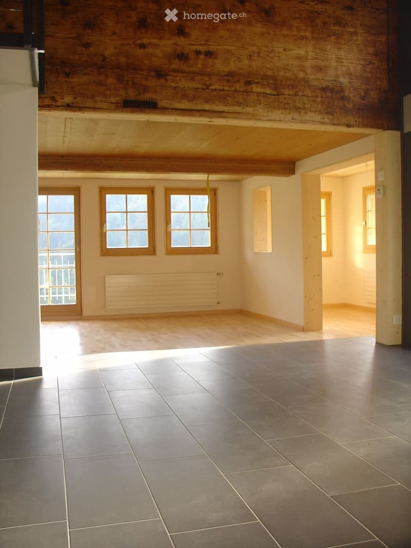 Empty room with wooden beams on the ceiling, large windows, and tiled floor. No furniture or equipment present.
