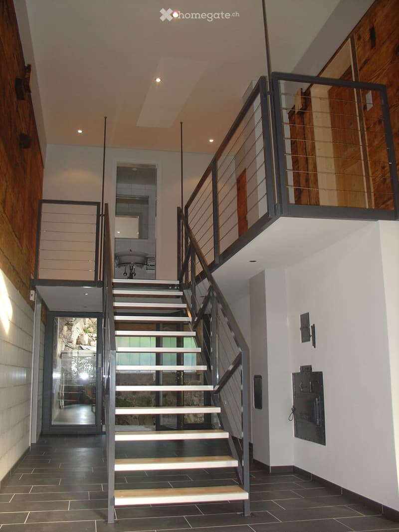 Modern interior staircase with wooden steps, metal railing, and glass panels, leading to a room with a sink visible in the background.