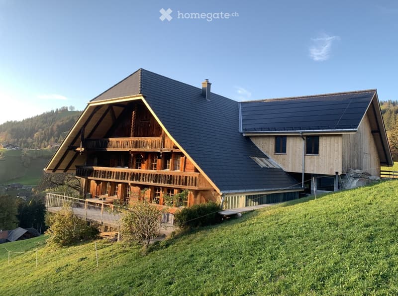 Two-story wooden house with a dark roof, large windows, and a wooden balcony overlooking a grassy hillside.