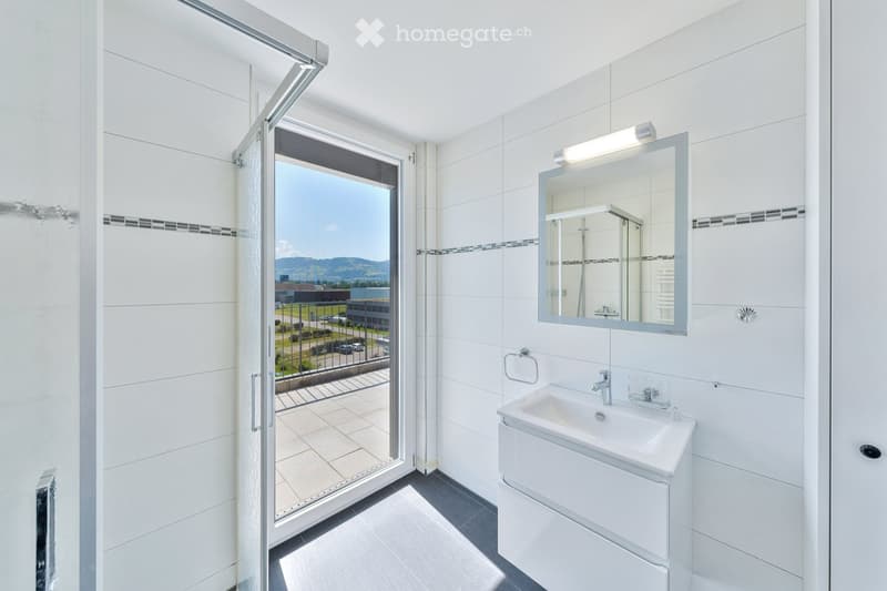 Modern bathroom with white tiles, a sink, a mirror, and a view of the outdoor area through a glass door.