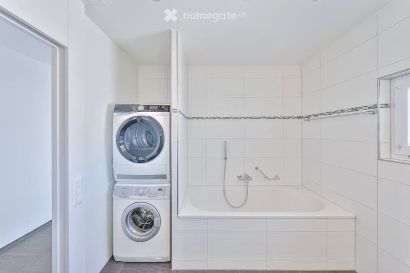 Modern bathroom with white tiled walls, a bathtub, and stacked washing machine and dryer unit.
