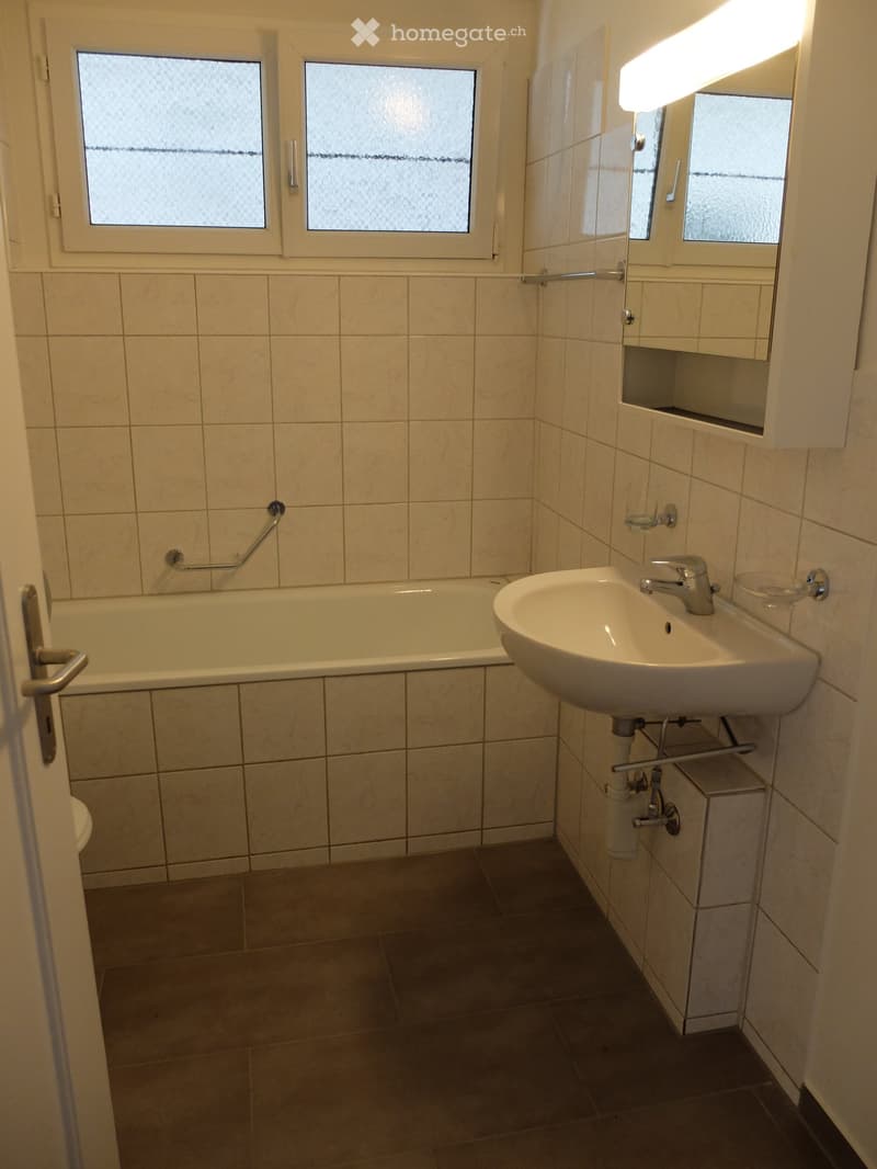 Bathroom with a bathtub, sink, mirror, and cabinet. The room features tiled walls and a window with frosted glass.