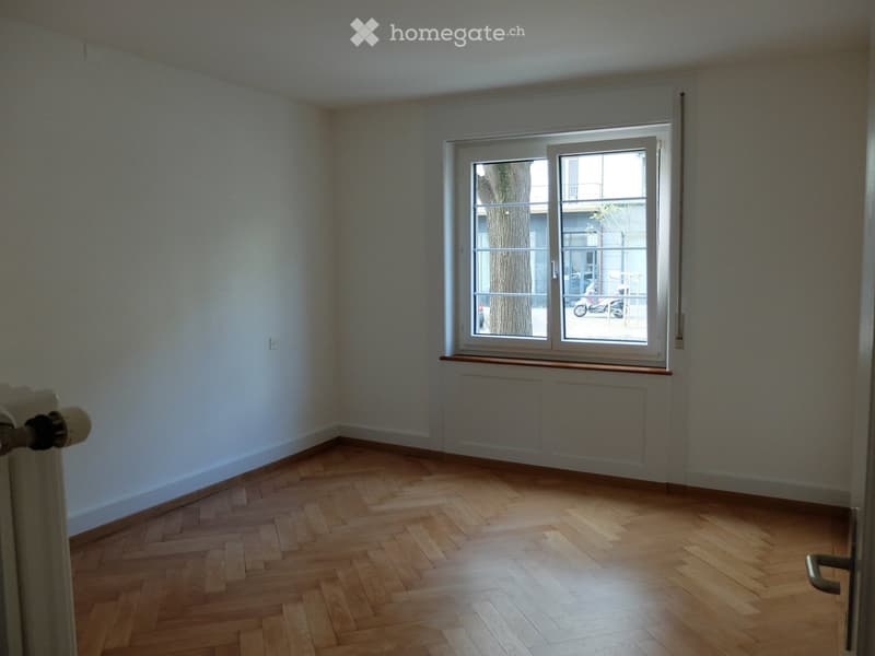 Empty room with wooden herringbone flooring, white walls, and a large window with a radiator below it.