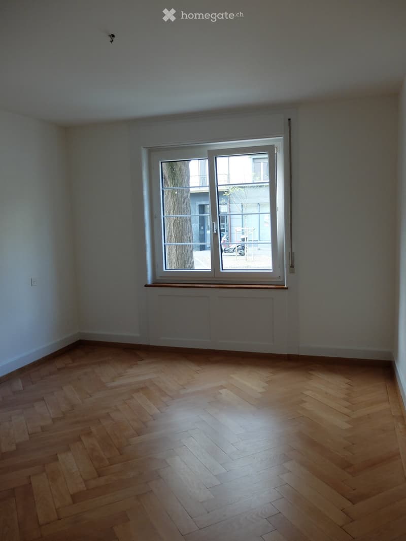 Empty room with wooden herringbone flooring, white walls, and a large window with a view of a tree and balcony area.