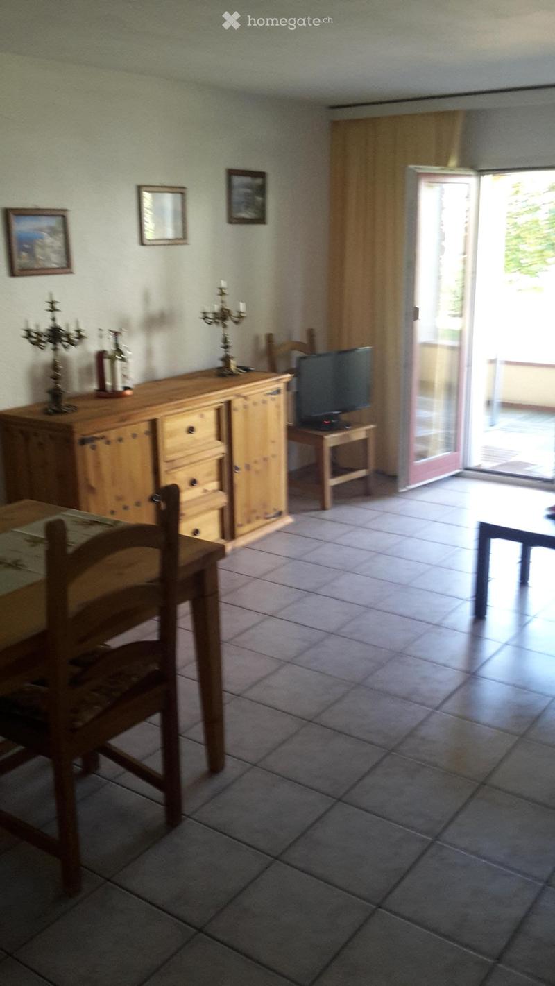 A furnished room with tiled flooring, a wooden chair, a small table, a wooden cabinet with drawers, a television on a stand, and candle holders on the cabinet.