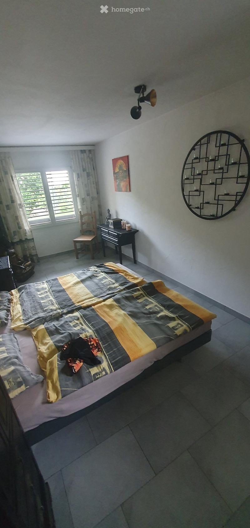Bedroom with a double bed, nightstand, chair, mirror, and painting on the wall. The bed has a patterned duvet and pillows.