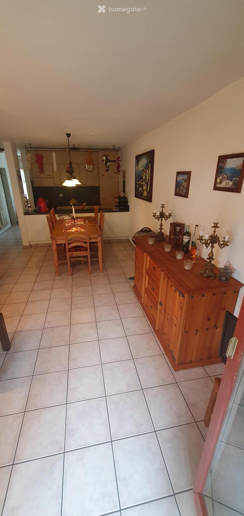 Dining area with a wooden dining table, chairs, a wooden cabinet with drawers, and decorative items including candle holders and picture frames. The room has tiled flooring and is connected to a kitchen area visible in the background.