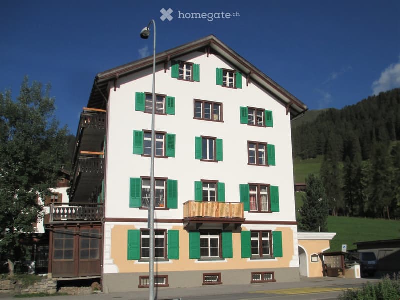A two-story house with white walls, green shutters, and wooden balconies, situated in a scenic, mountainous area.