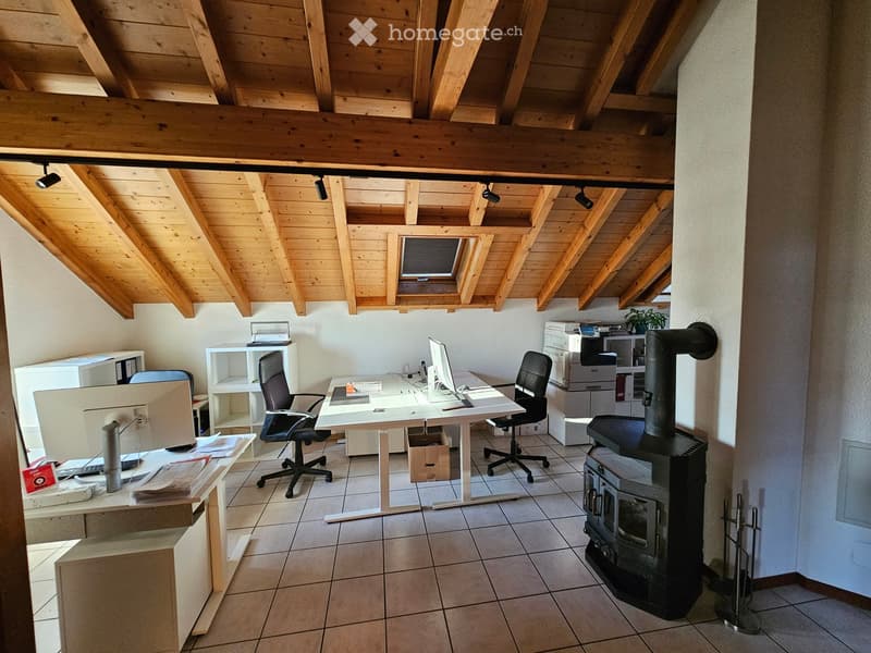 Office space with wooden ceiling beams, tiled floor, desk with computer, chairs, printer, and wood stove.