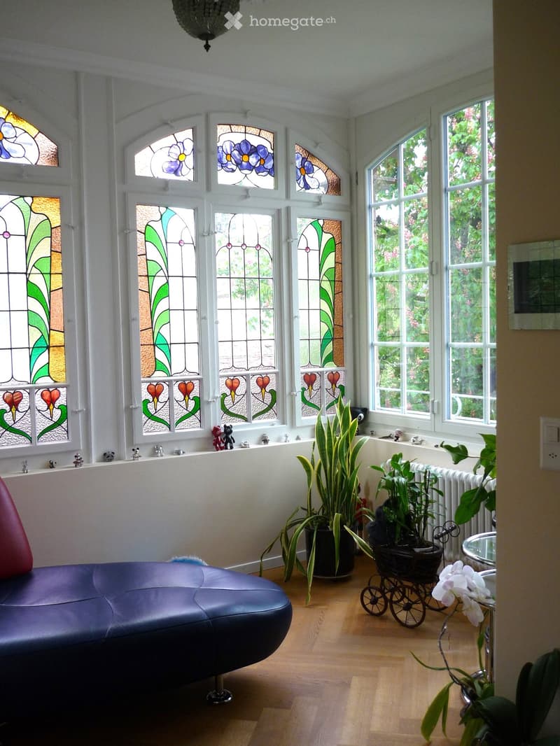 A modern living room with a blue leather sofa, wooden floor, large windows with stained glass, potted plants, and decorative toys on the windowsill.