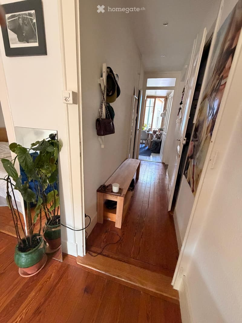 A narrow hallway with wooden flooring, white walls, and a small bench with a cup on it. A coat rack with hats and a bag is visible, and a potted plant is placed near the wall on the left side.