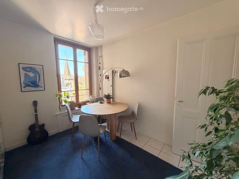 Bright room with a round wooden table, four chairs, a guitar, a floor lamp, potted plants, and a picture frame on the wall.