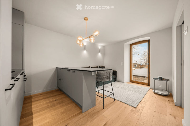 Open-plan living and dining area with modern kitchen island, bar stool, sofa, coffee table, side table, and a hanging chandelier. The space features light wood flooring and large windows with a view of the street.