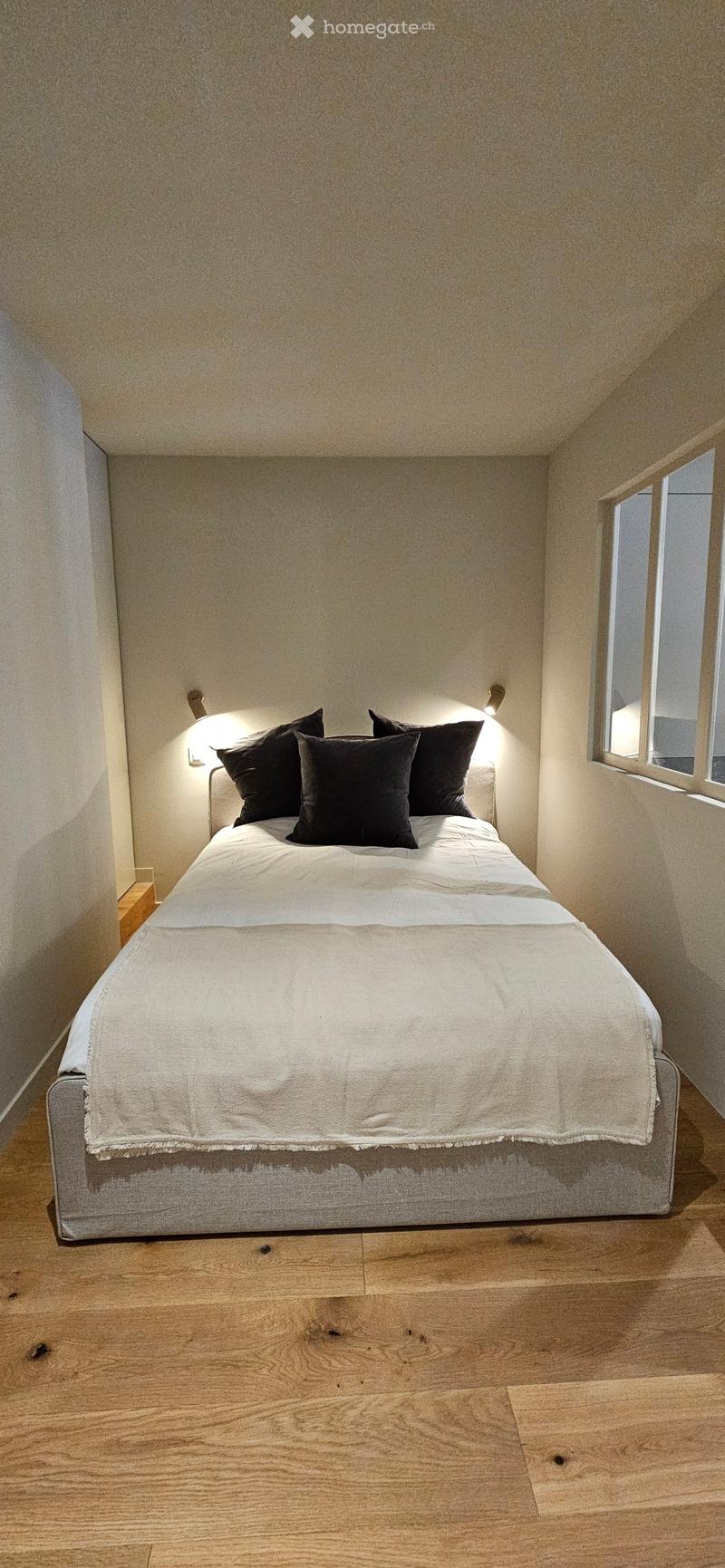 A modern bedroom with a neatly made bed, white bedding, black pillows, and a wooden nightstand. The room features light-colored walls and wooden flooring.