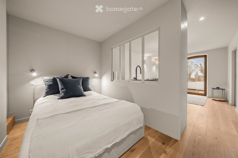 Modern bedroom with a large bed, dark blue pillows, wall-mounted lamps, and a view into an adjacent kitchen area with a modern faucet and mirror. A small table and chair are visible near the doorway.