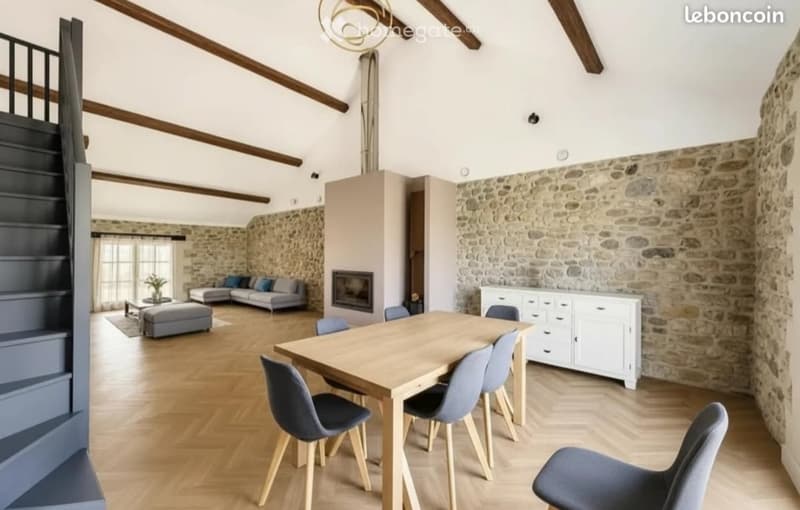 Open-plan living and dining area with exposed wooden beams, stone walls, fireplace, grey sofa, coffee table, ottoman, dining table with chairs, and white cabinet.