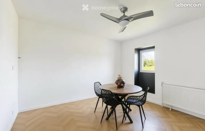 A modern, minimalist dining area with a round wooden table, four black wire chairs, a ceiling fan, and a radiator. The room features light-colored walls and a herringbone wooden floor.