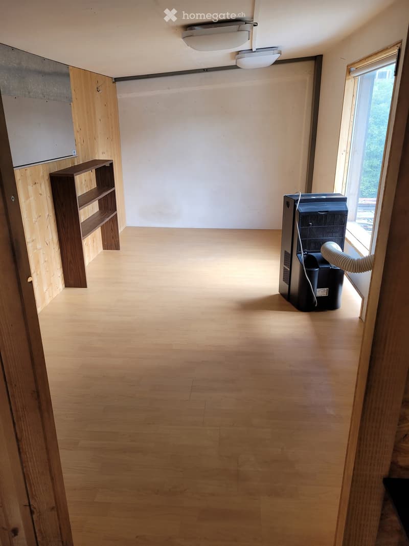 Empty room with light wooden flooring, white walls, and a large window letting in natural light. A small wooden shelf is visible on the left side, and an air conditioning unit is placed near the window on the right.