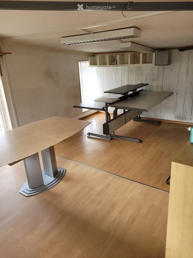 A modern, unfurnished room with a large wooden table and a grey adjustable standing desk, featuring a light wooden floor and white walls.